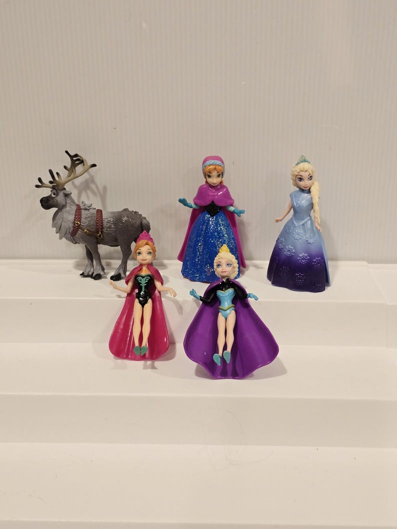 May include: A collection of miniature figurines featuring characters from the animated film "Frozen." The set includes Elsa, Anna, and other characters, with detailed costumes and accessories. The figures are displayed on a white stepped surface.