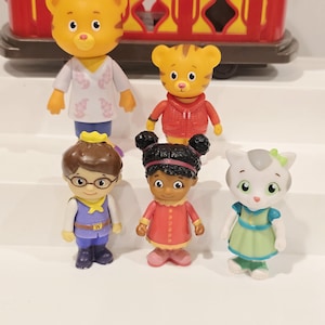 May include: A collection of six Daniel Tiger's Neighborhood figurines. The figurines include Daniel Tiger, Katerina Kittycat, O the Owl, Prince Wednesday, and Miss Elaina. The figures are brightly colored and made of plastic. A red and yellow trolley is in the background.