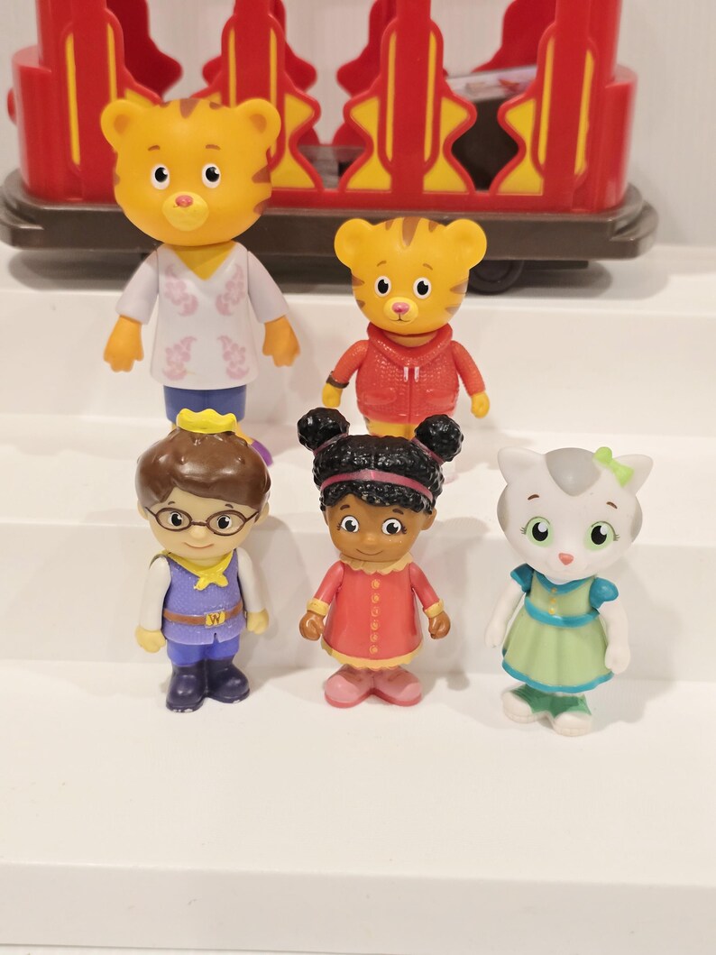 May include: A collection of six Daniel Tiger's Neighborhood figurines. The figurines include Daniel Tiger, Katerina Kittycat, O the Owl, Prince Wednesday, and Miss Elaina. The figures are brightly colored and made of plastic. A red and yellow trolley is in the background.