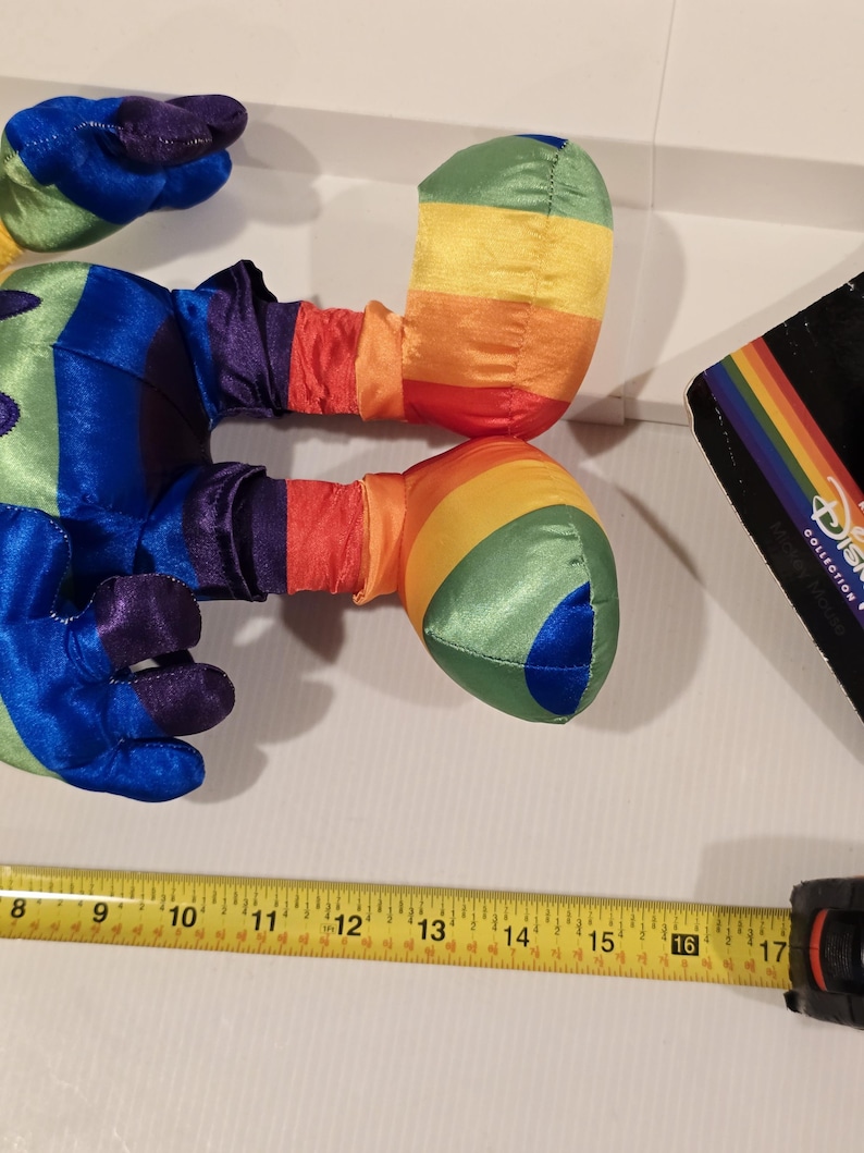 May include: A colorful plush toy with rainbow-striped arms and legs, and blue, purple, and green accents. The toy is next to a measuring tape showing 16 inches. A Disney Collection tag is visible.
