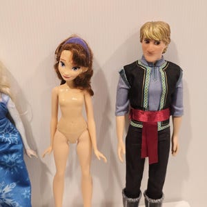 May include: Two dolls are shown. One has brown hair and a purple headband. The other doll has blonde hair and wears a black vest with blue and green trim, black trousers, a red sash, and black boots.