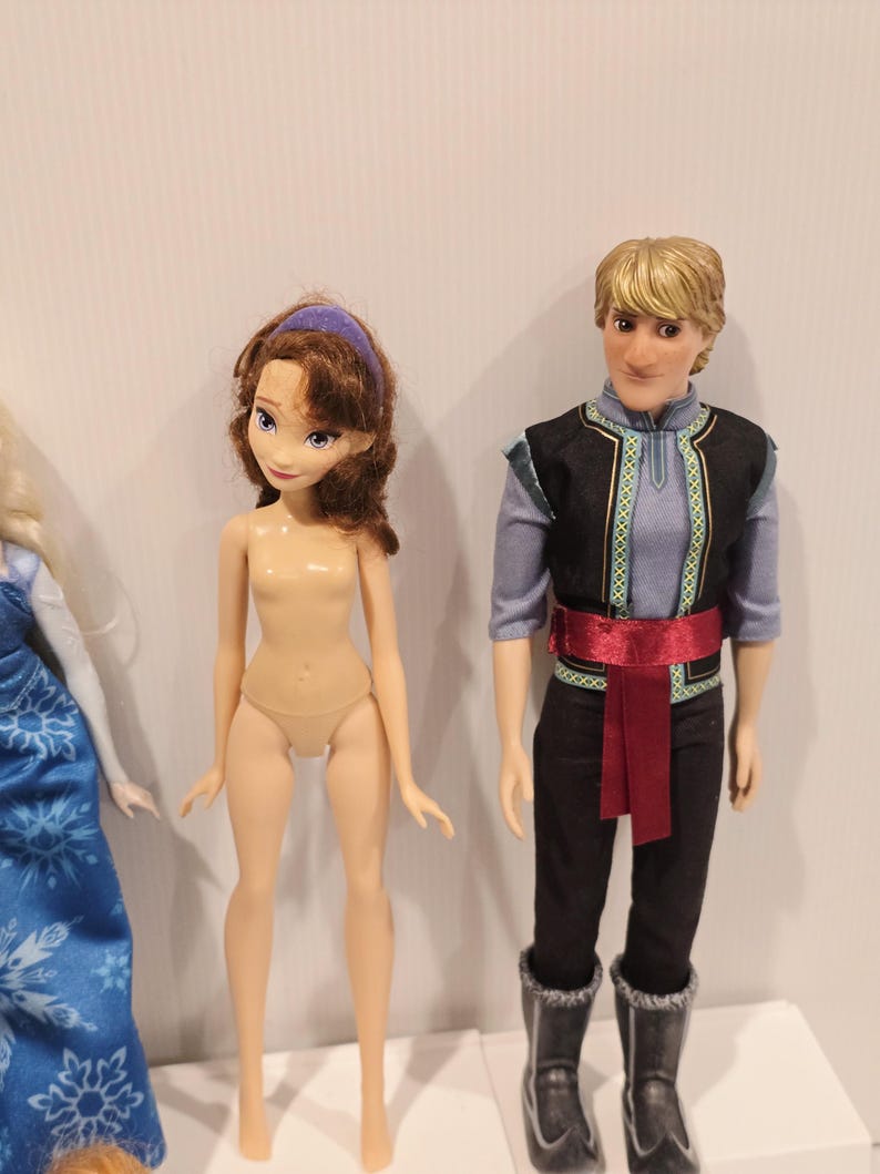 May include: Two dolls are shown. One has brown hair and a purple headband. The other doll has blonde hair and wears a black vest with blue and green trim, black trousers, a red sash, and black boots.