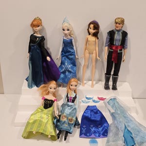 May include: A selection of Disney Frozen dolls and accessories. The dolls feature Elsa, Anna, and Kristoff, each in their iconic outfits. Additional clothing and accessories are included, such as a blue skirt and a translucent blue gown.