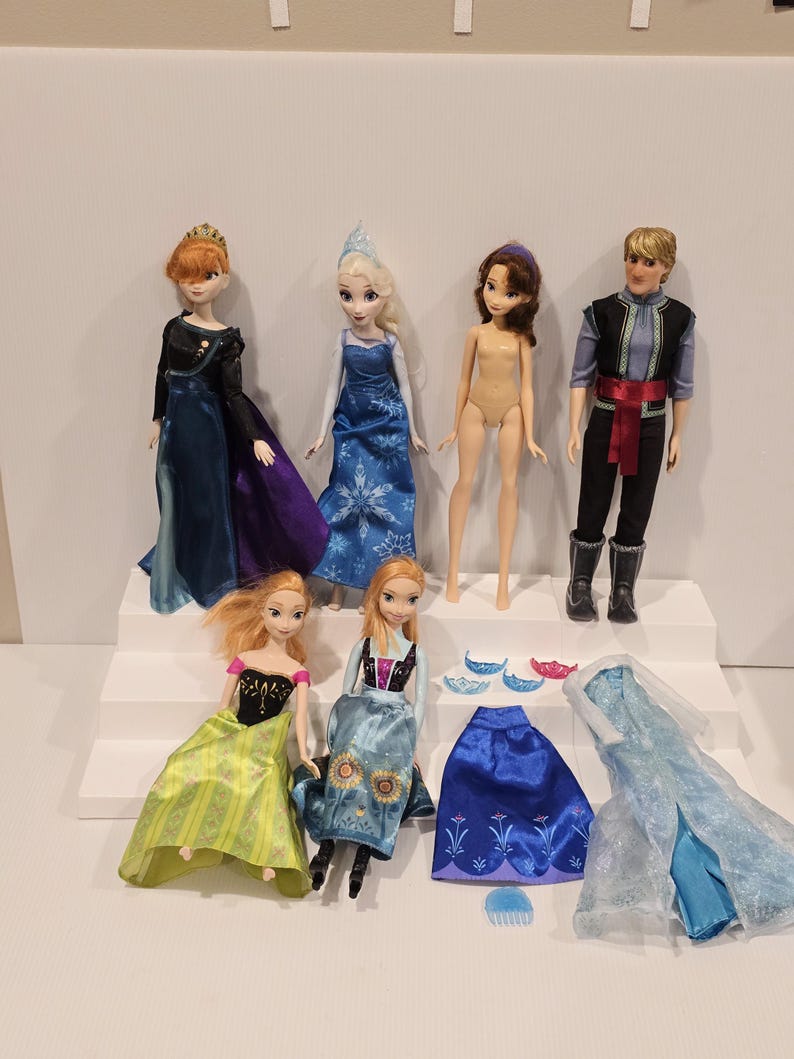 May include: A selection of Disney Frozen dolls and accessories. The dolls feature Elsa, Anna, and Kristoff, each in their iconic outfits. Additional clothing and accessories are included, such as a blue skirt and a translucent blue gown.