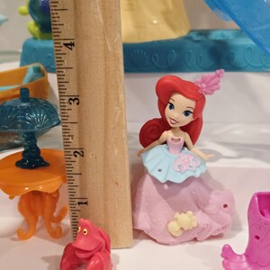 K&ouml;nnte beinhalten: Ariel-Puppe aus Arielle, die Meerjungfrau, mit einem rosa Kleid mit hellblauem Oberteil und rotem Haar. Eine rote Krabbenfigur und ein orangefarbener Tisch sind ebenfalls zu sehen, zusammen mit anderem Spielzeugzubeh&ouml;r. Das Holzlineal zeigt die Ma&szlig;e in Zentimetern.