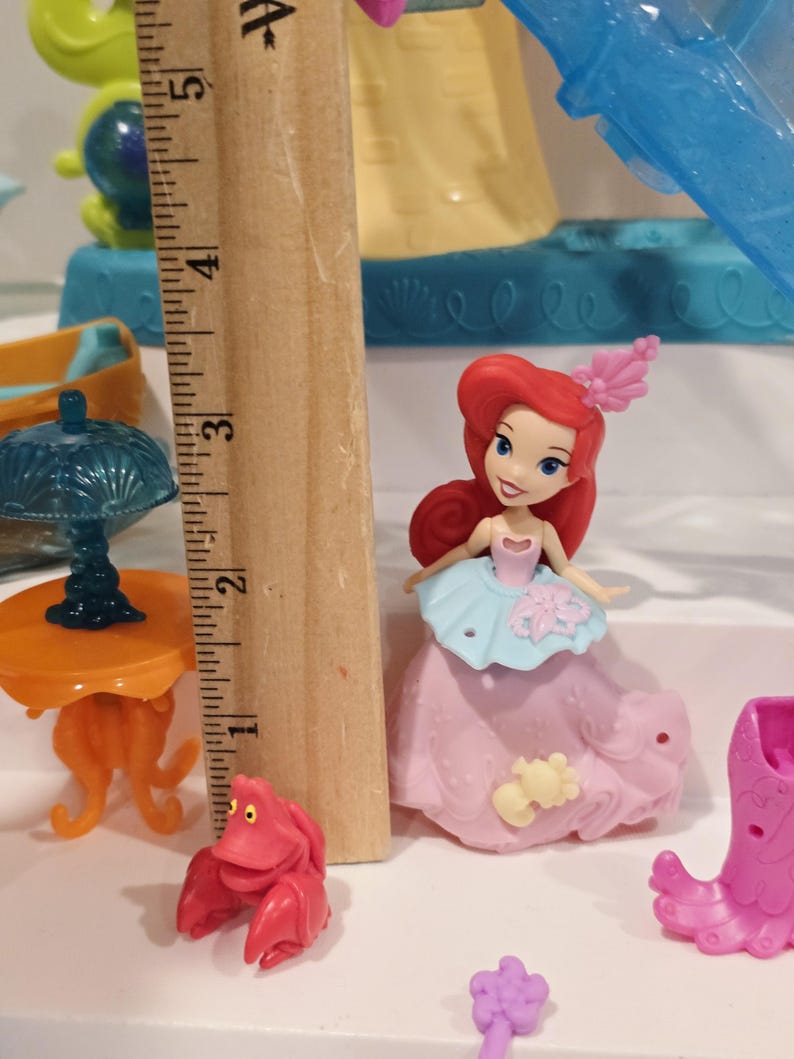 K&ouml;nnte beinhalten: Ariel-Puppe aus Arielle, die Meerjungfrau, mit einem rosa Kleid mit hellblauem Oberteil und rotem Haar. Eine rote Krabbenfigur und ein orangefarbener Tisch sind ebenfalls zu sehen, zusammen mit anderem Spielzeugzubeh&ouml;r. Das Holzlineal zeigt die Ma&szlig;e in Zentimetern.