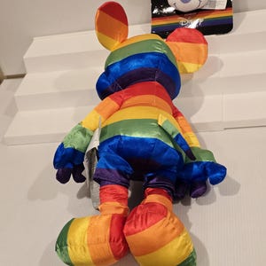 May include: A rainbow-striped Mickey Mouse plush toy with matching ears, arms, legs, and feet. The plush toy is made of a soft, shiny material. A smaller Mickey Mouse head is attached to a black tag in the background.
