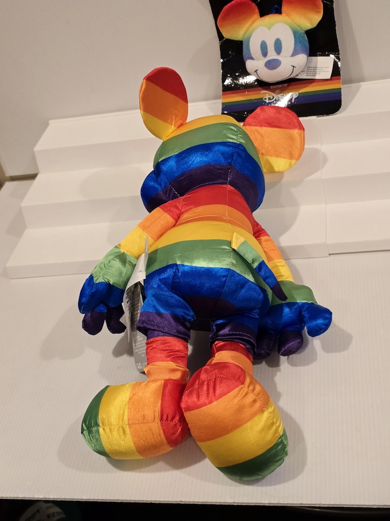 May include: A rainbow-striped Mickey Mouse plush toy with matching ears, arms, legs, and feet. The plush toy is made of a soft, shiny material. A smaller Mickey Mouse head is attached to a black tag in the background.