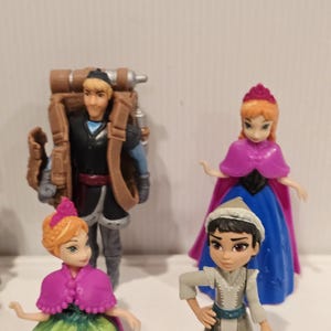 May include: Four miniature figurines from the animated film Frozen. The figures include Anna and Kristoff, with Anna wearing a pink cape and a green and gold dress. Kristoff has a brown backpack. Another Anna and a character in a white and gray outfit are also present.