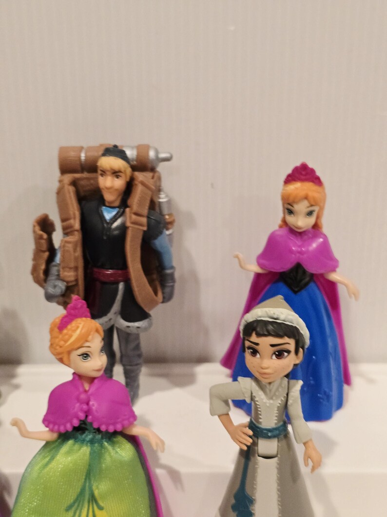 May include: Four miniature figurines from the animated film Frozen. The figures include Anna and Kristoff, with Anna wearing a pink cape and a green and gold dress. Kristoff has a brown backpack. Another Anna and a character in a white and gray outfit are also present.