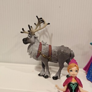 May include: A grey toy reindeer with large antlers and a red harness stands next to a doll of a character with a pink cape and dress. The reindeer has a detailed fur texture and is posed in a standing position.