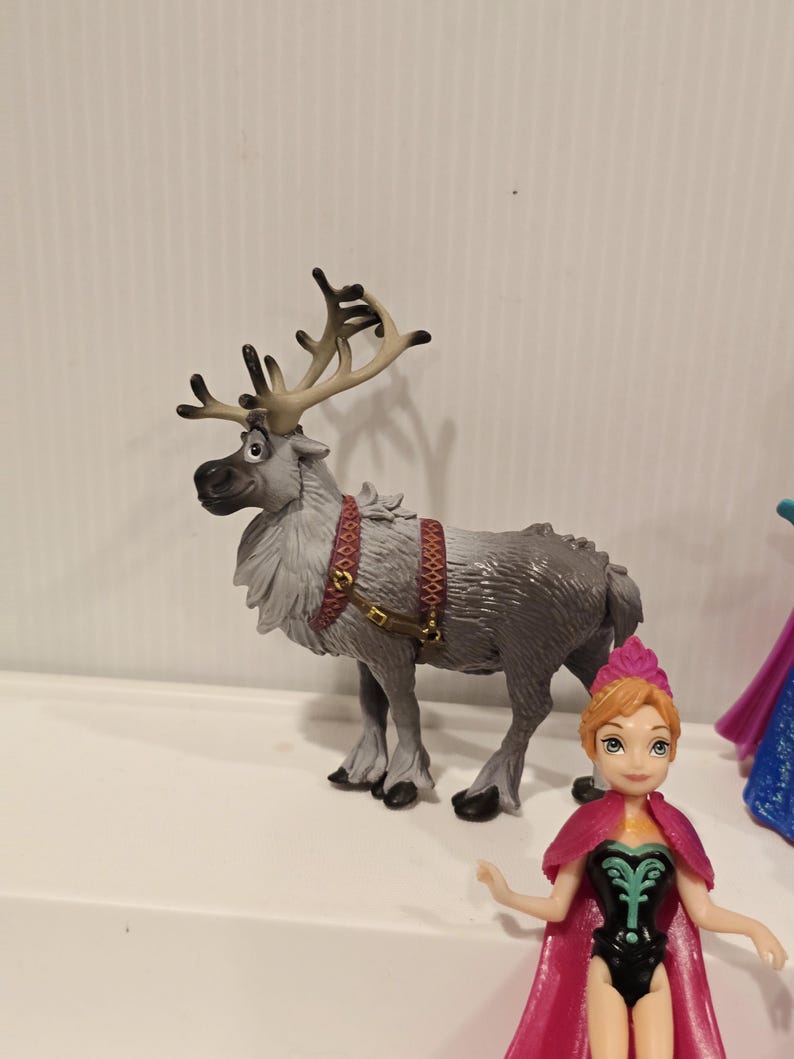 May include: A grey toy reindeer with large antlers and a red harness stands next to a doll of a character with a pink cape and dress. The reindeer has a detailed fur texture and is posed in a standing position.