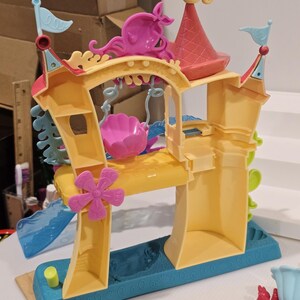 May include: A colorful plastic playset resembling an underwater castle. The playset features a yellow base with blue accents, pink and yellow towers, and a pink seashell-shaped swing. A seahorse figure tops the red roof.