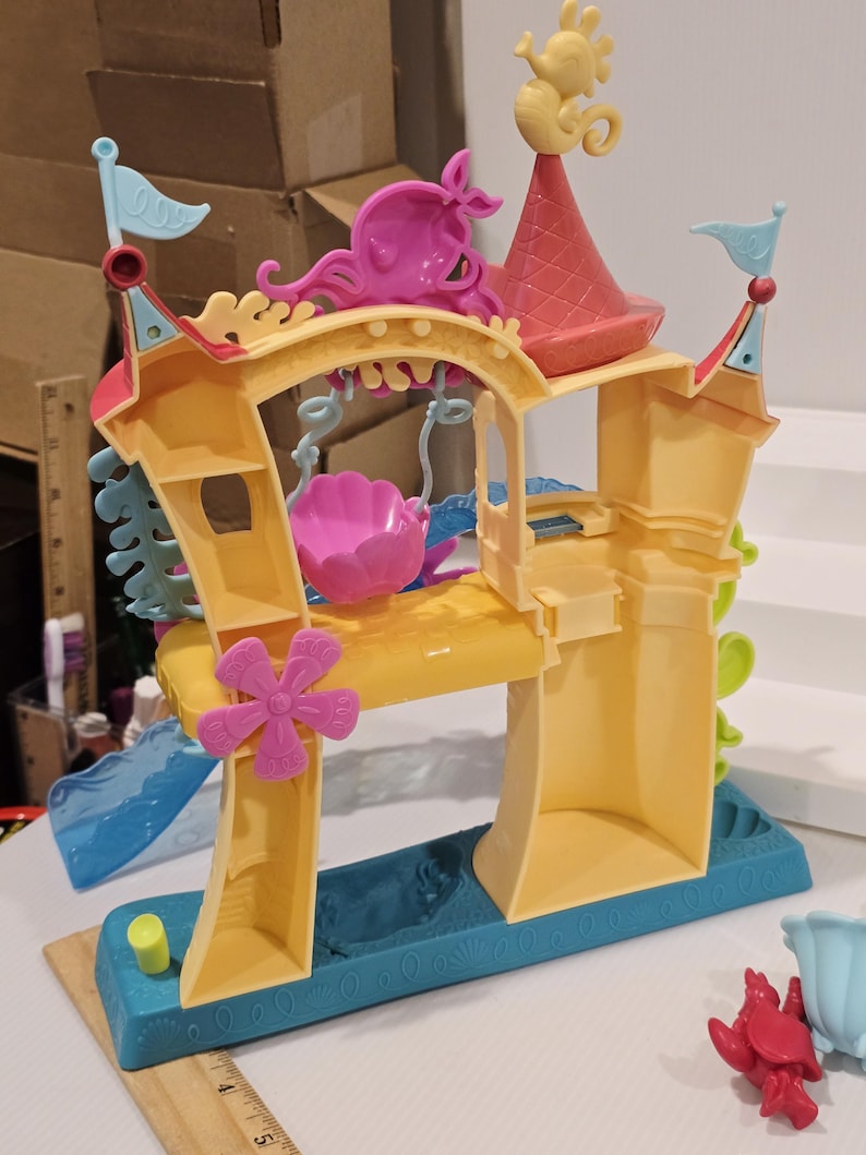May include: A colorful plastic playset resembling an underwater castle. The playset features a yellow base with blue accents, pink and yellow towers, and a pink seashell-shaped swing. A seahorse figure tops the red roof.