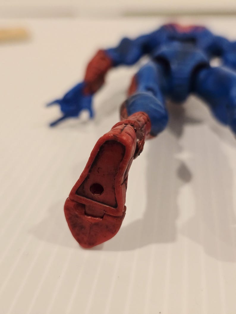 Pode incluir: Uma figura de a&ccedil;&atilde;o azul e vermelha de um super-her&oacute;i, deitada. A figura tem um fato azul com detalhes vermelhos nos bra&ccedil;os e nos p&eacute;s. O p&eacute; da figura est&aacute; em primeiro plano, mostrando a bota vermelha com um buraco.