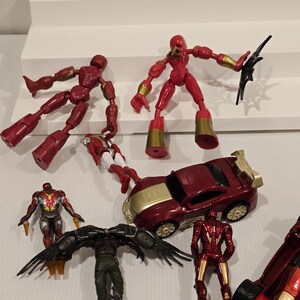 May include: A collection of red and gold action figures and a red and gold toy car are displayed. The figures include articulated characters with gold accents and a winged figure. The toy car has gold detailing.