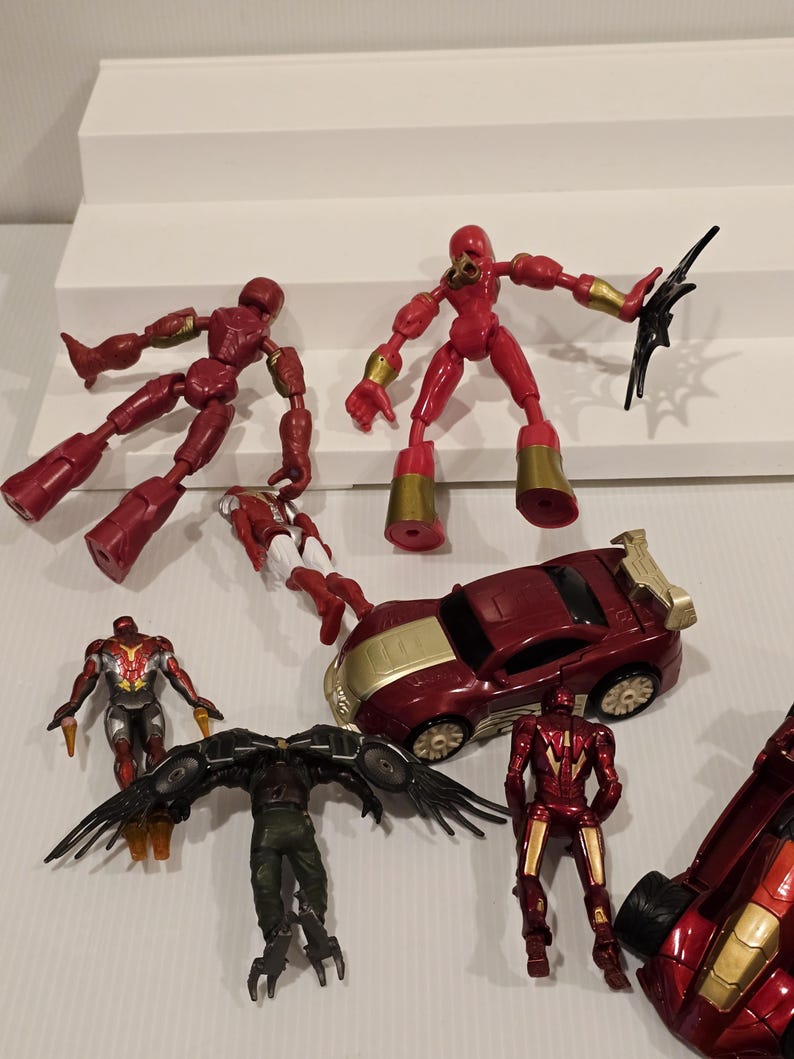 May include: A collection of red and gold action figures and a red and gold toy car are displayed. The figures include articulated characters with gold accents and a winged figure. The toy car has gold detailing.