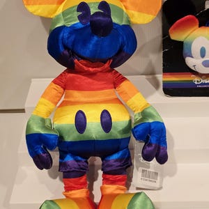 May include: A plush Mickey Mouse toy in a rainbow color scheme. The toy features a yellow head, orange ears, and a red, orange, yellow, green, blue, and purple striped body. The toy has black eyes and a black nose. A tag with a barcode is attached.