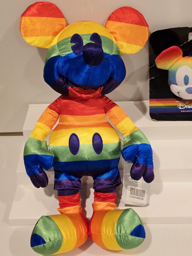 May include: A plush Mickey Mouse toy in a rainbow color scheme. The toy features a yellow head, orange ears, and a red, orange, yellow, green, blue, and purple striped body. The toy has black eyes and a black nose. A tag with a barcode is attached.