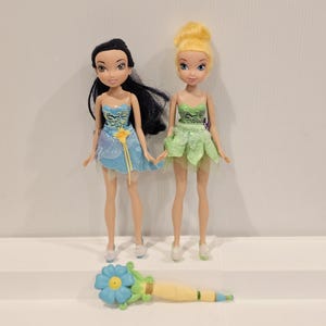 May include: Two dolls, one with black hair and a blue dress, the other with blonde hair and a green dress, stand side-by-side. A blue and yellow flower-shaped wand lies in front of them. The dolls are on a white surface.