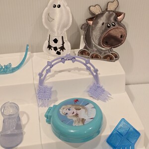 K&ouml;nnte beinhalten: Eine Sammlung von Spielzeug und Zubeh&ouml;r zum Thema Frozen. Enth&auml;lt eine hellblaue Kompaktpuder mit einem Bild von Elsa, ein wei&szlig;es Olaf-Pl&uuml;schtier, ein graues Rentier-Pl&uuml;schtier, einen blauen Kamm und weiteres kleines, hellblaues Zubeh&ouml;r.