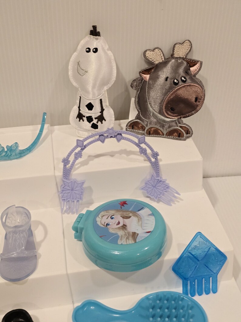 K&ouml;nnte beinhalten: Eine Sammlung von Spielzeug und Zubeh&ouml;r zum Thema Frozen. Enth&auml;lt eine hellblaue Kompaktpuder mit einem Bild von Elsa, ein wei&szlig;es Olaf-Pl&uuml;schtier, ein graues Rentier-Pl&uuml;schtier, einen blauen Kamm und weiteres kleines, hellblaues Zubeh&ouml;r.