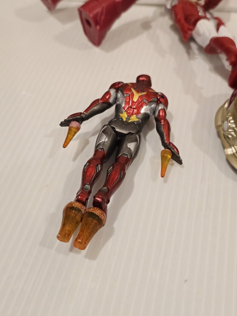 May include: A red, silver, and gold action figure of a superhero in a flying pose. The figure has a detailed design with a metallic finish. The figure has orange cone-shaped elements coming from the hands and feet.