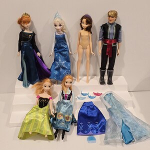 May include: A collection of Disney Frozen dolls and accessories. The dolls include Elsa, Anna, Kristoff, and other characters. Accessories include dresses, capes, and tiaras. The dolls are posed on a white stepped platform.