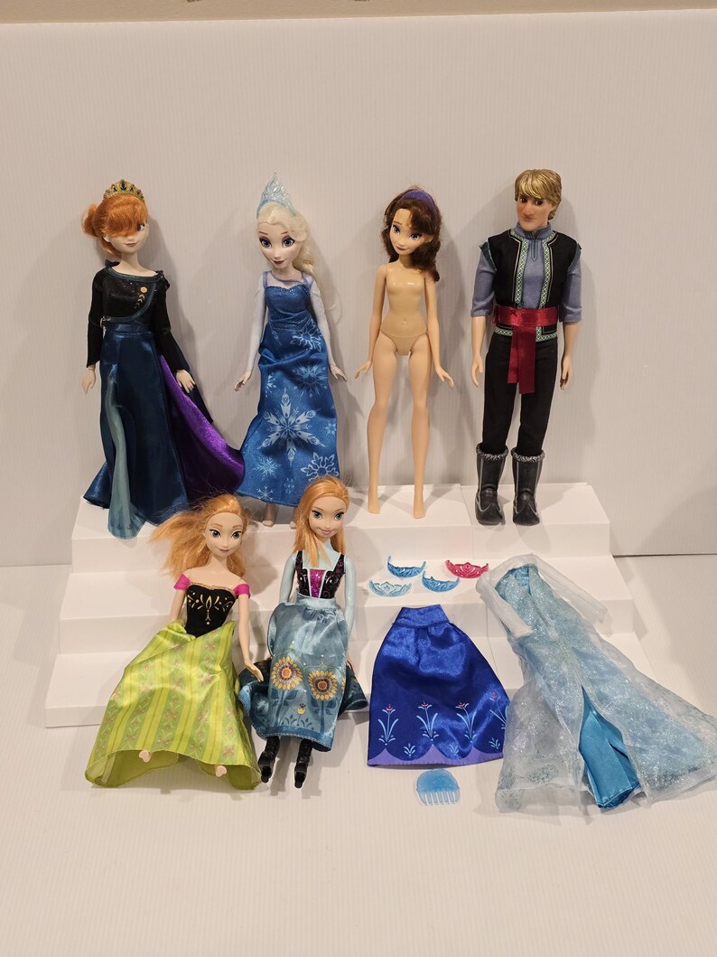 May include: A collection of Disney Frozen dolls and accessories. The dolls include Elsa, Anna, Kristoff, and other characters. Accessories include dresses, capes, and tiaras. The dolls are posed on a white stepped platform.