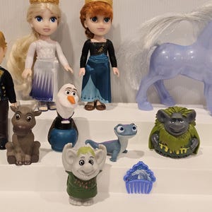 May include: A collection of Frozen-themed figurines, including Elsa and Anna dolls with golden crowns, Olaf the snowman, a reindeer, a horse, and other characters. The dolls are dressed in their signature outfits, with vibrant colors and detailed designs.