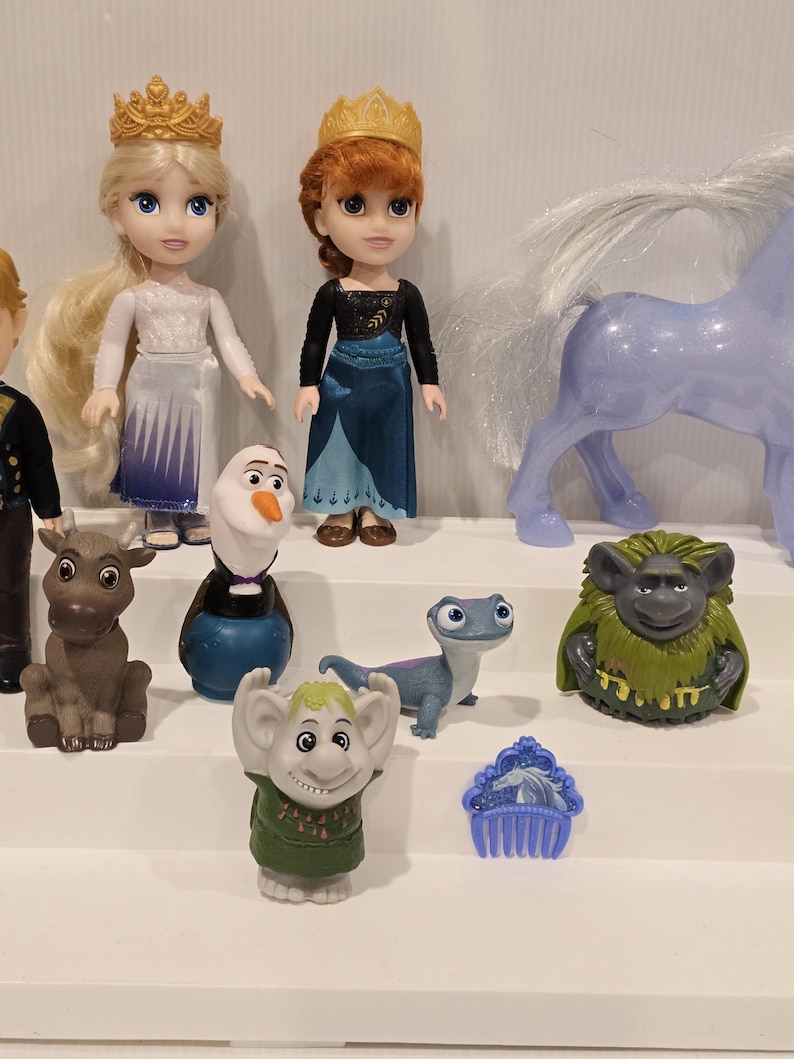 May include: A collection of Frozen-themed figurines, including Elsa and Anna dolls with golden crowns, Olaf the snowman, a reindeer, a horse, and other characters. The dolls are dressed in their signature outfits, with vibrant colors and detailed designs.