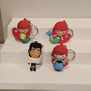 May include: Four character keychains featuring characters from a popular animated film. The keychains include two mermaids with red hair and blue and green tails, a male character in a white and blue outfit, and a mermaid in a blue dress.