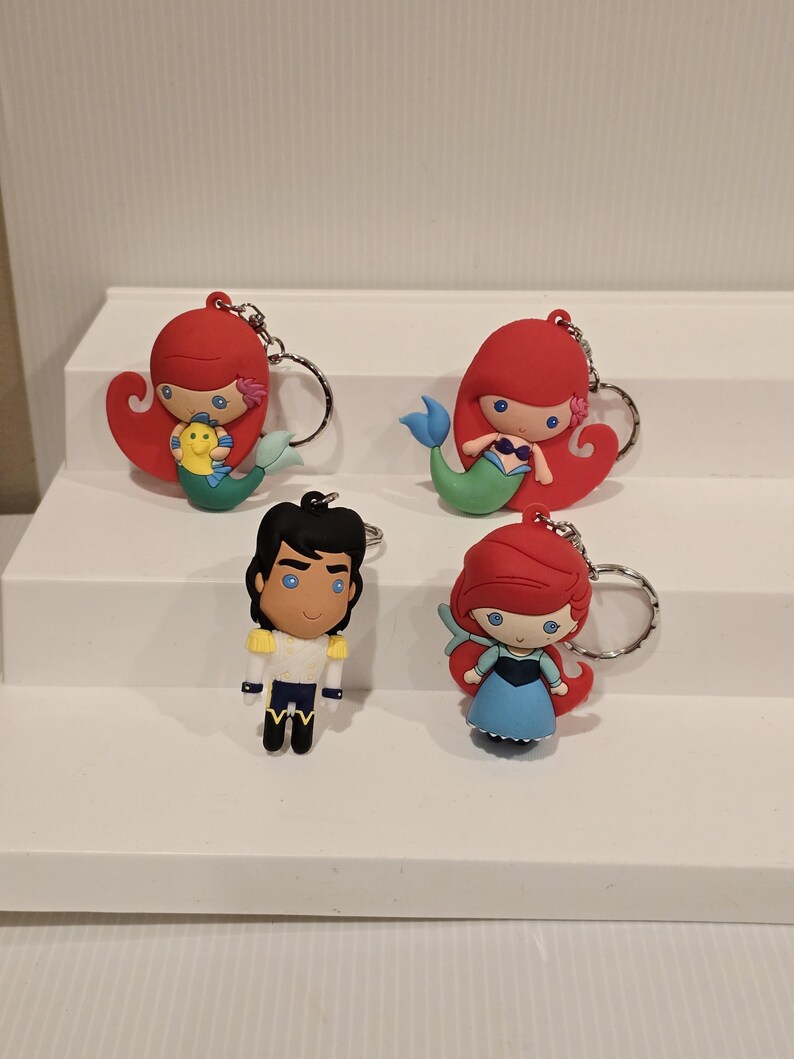 May include: Four character keychains featuring characters from a popular animated film. The keychains include two mermaids with red hair and blue and green tails, a male character in a white and blue outfit, and a mermaid in a blue dress.