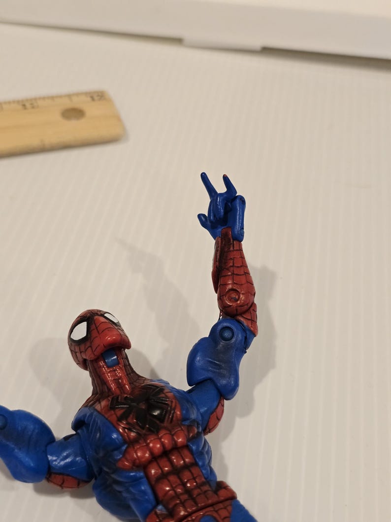 Pode incluir: Figura de a&ccedil;&atilde;o do Homem-Aranha em pose din&acirc;mica, com traje vermelho e azul e detalhes de teia de aranha. O bra&ccedil;o est&aacute; levantado, a m&atilde;o faz um gesto. Olhos brancos e emblema de aranha preta no peito.