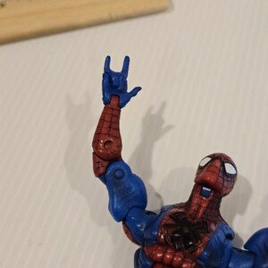 Pode incluir: Figura de a&ccedil;&atilde;o do Homem-Aranha em pose din&acirc;mica, com um traje vermelho e azul com detalhes de teia. O bra&ccedil;o da figura est&aacute; levantado, mostrando um gesto com a m&atilde;o. Uma r&eacute;gua de madeira est&aacute; no fundo.