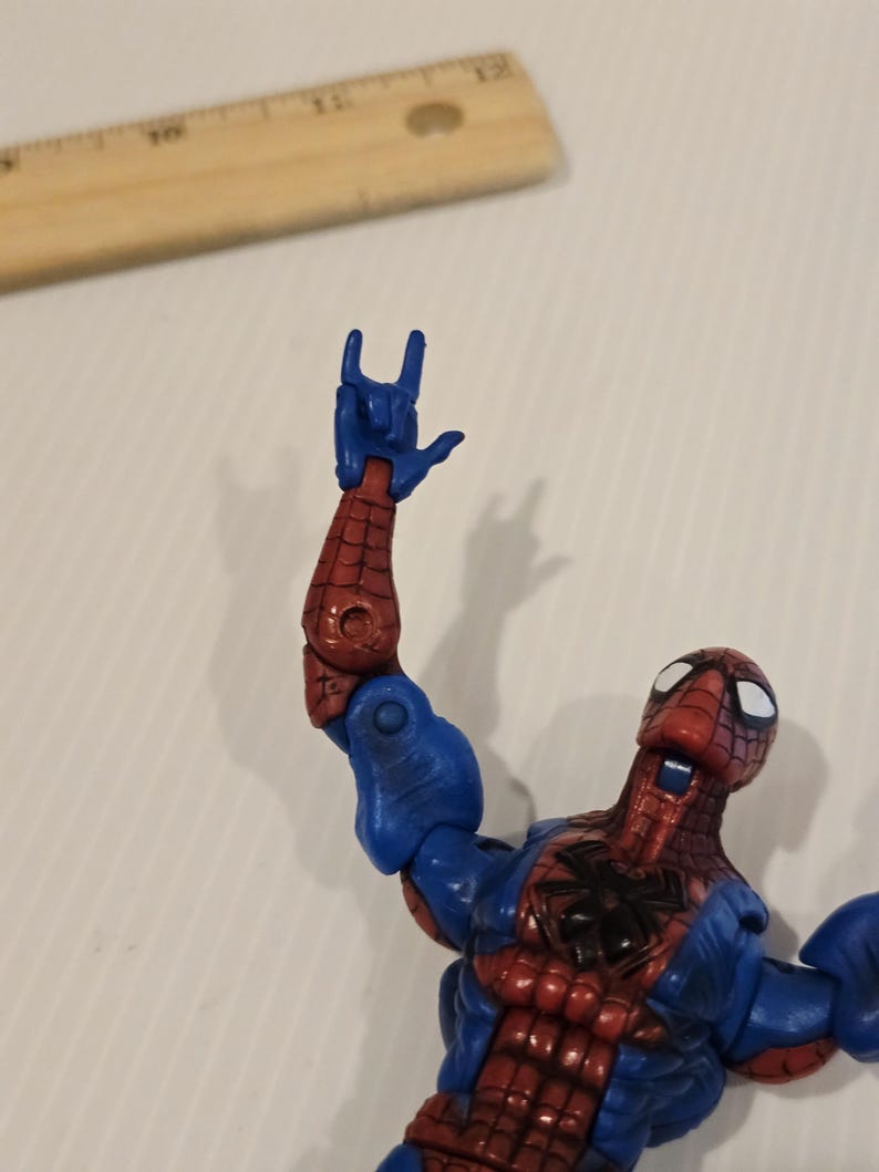 Pode incluir: Figura de a&ccedil;&atilde;o do Homem-Aranha em pose din&acirc;mica, com um traje vermelho e azul com detalhes de teia. O bra&ccedil;o da figura est&aacute; levantado, mostrando um gesto com a m&atilde;o. Uma r&eacute;gua de madeira est&aacute; no fundo.