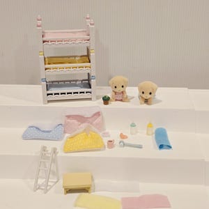 Puede incluir: Un juego de guarder&iacute;a en miniatura para casa de mu&ntilde;ecas con una litera, dos figuras de cachorros y varios accesorios. Incluye una mesita, una escalera, mantas, biberones y un pa&ntilde;o. La litera tiene detalles en rosa, amarillo y azul.