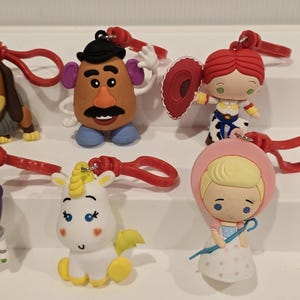 May include: A selection of character keyrings from a popular animated film series. The keyrings include a potato head, a cowgirl, a unicorn, and a girl in a pink bonnet. Each keyring has a red clasp and is brightly coloured.