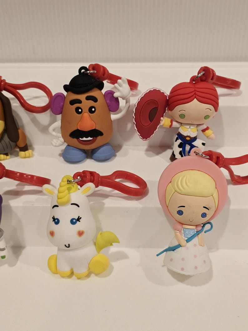 May include: A selection of character keyrings from a popular animated film series. The keyrings include a potato head, a cowgirl, a unicorn, and a girl in a pink bonnet. Each keyring has a red clasp and is brightly coloured.