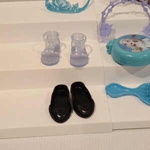 K&ouml;nnte beinhalten: Eine Sammlung von Kinderspielzeug, darunter ein hellblaues Diadem, zwei Paar Schuhe und ein blauer Kompaktpuder mit einem Charakterbild. Die Schuhe umfassen schwarze Ballerinas und zwei transparente Sandalen.