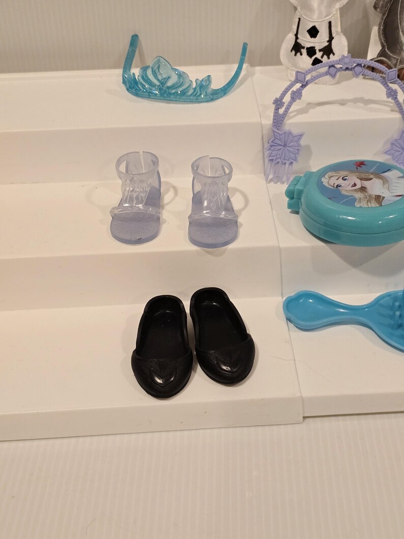 K&ouml;nnte beinhalten: Eine Sammlung von Kinderspielzeug, darunter ein hellblaues Diadem, zwei Paar Schuhe und ein blauer Kompaktpuder mit einem Charakterbild. Die Schuhe umfassen schwarze Ballerinas und zwei transparente Sandalen.