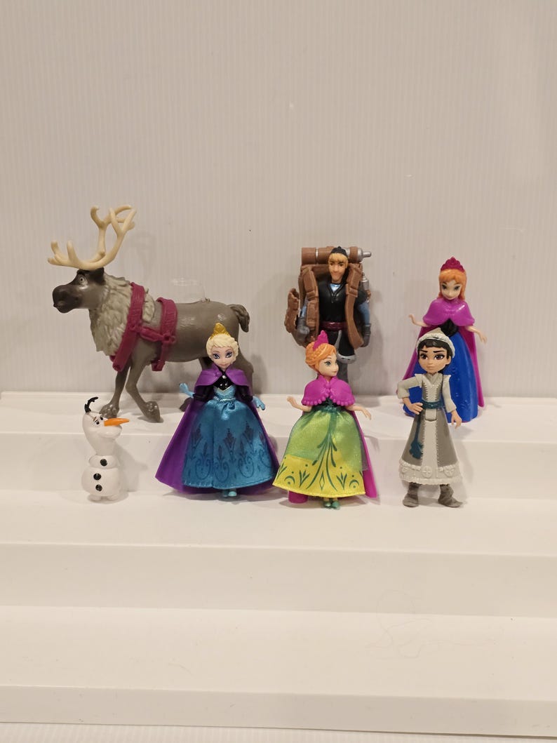 May include: A collection of miniature figurines featuring characters from the animated film "Frozen." The set includes Elsa, Anna, Kristoff, Olaf, and Sven. The figurines are dressed in their signature outfits, with vibrant colors and detailed designs.