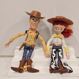 May include: Two character dolls, Woody and Jessie, from the Toy Story franchise. Woody wears a brown cowboy hat, yellow plaid shirt, blue jeans, and brown boots. Jessie wears a brown hat, white shirt, blue jeans with cow print chaps, and brown boots.