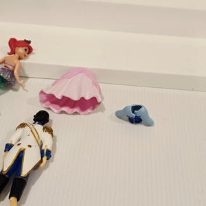 K&ouml;nnte beinhalten: Eine Sammlung von Miniaturfiguren aus einem Kinderanimationsfilm. Die Figuren umfassen eine Meerjungfrau, einen Prinzen, eine Krabbe und weiteres Zubeh&ouml;r. Die Meerjungfrau hat rotes Haar und einen lila-gr&uuml;nen Schwanz. Der Prinz tr&auml;gt eine wei&szlig;e Jacke mit goldenen Verzierungen.