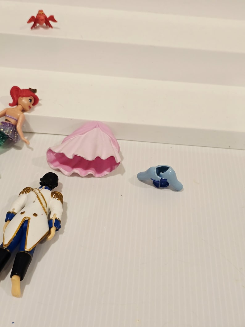 K&ouml;nnte beinhalten: Eine Sammlung von Miniaturfiguren aus einem Kinderanimationsfilm. Die Figuren umfassen eine Meerjungfrau, einen Prinzen, eine Krabbe und weiteres Zubeh&ouml;r. Die Meerjungfrau hat rotes Haar und einen lila-gr&uuml;nen Schwanz. Der Prinz tr&auml;gt eine wei&szlig;e Jacke mit goldenen Verzierungen.