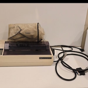 May include: A beige Tandy printer with a black print head and control panel. A clear plastic cover with the word "TANDY" is on top. A black cable is connected to the printer.