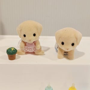 Puede incluir: Dos peque&ntilde;as figuras de cachorros de color canela claro con ojos negros. Un cachorro lleva un vestido rosa y el otro un babero azul. Una peque&ntilde;a planta verde en una maceta marr&oacute;n est&aacute; al lado del primer cachorro. Biberones en primer plano.
