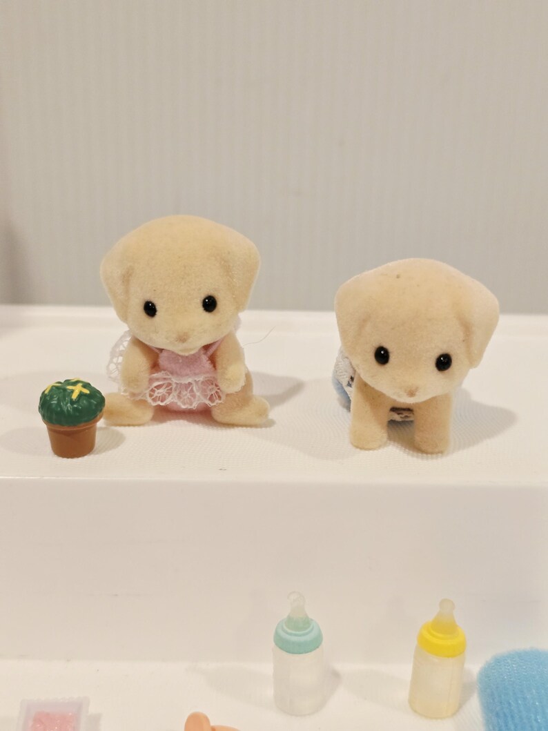 Puede incluir: Dos peque&ntilde;as figuras de cachorros de color canela claro con ojos negros. Un cachorro lleva un vestido rosa y el otro un babero azul. Una peque&ntilde;a planta verde en una maceta marr&oacute;n est&aacute; al lado del primer cachorro. Biberones en primer plano.