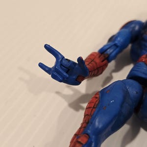 Pode incluir: Uma figura de a&ccedil;&atilde;o azul e vermelha de um super-her&oacute;i, com um gesto com a m&atilde;o. A figura tem um fato azul com detalhes vermelhos nos bra&ccedil;os e pernas. A figura est&aacute; numa pose din&acirc;mica, com uma m&atilde;o estendida.