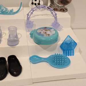 K&ouml;nnte beinhalten: Eine Sammlung von Kinderspielzeug und Zubeh&ouml;r, darunter eine blaue Haarb&uuml;rste, schwarze Schuhe und ein blauer Kompaktpuder mit einem Charakterbild. Weitere Artikel sind ein Diadem, ein klarer Schuh und ein blaues, eisgeformtes Accessoire.
